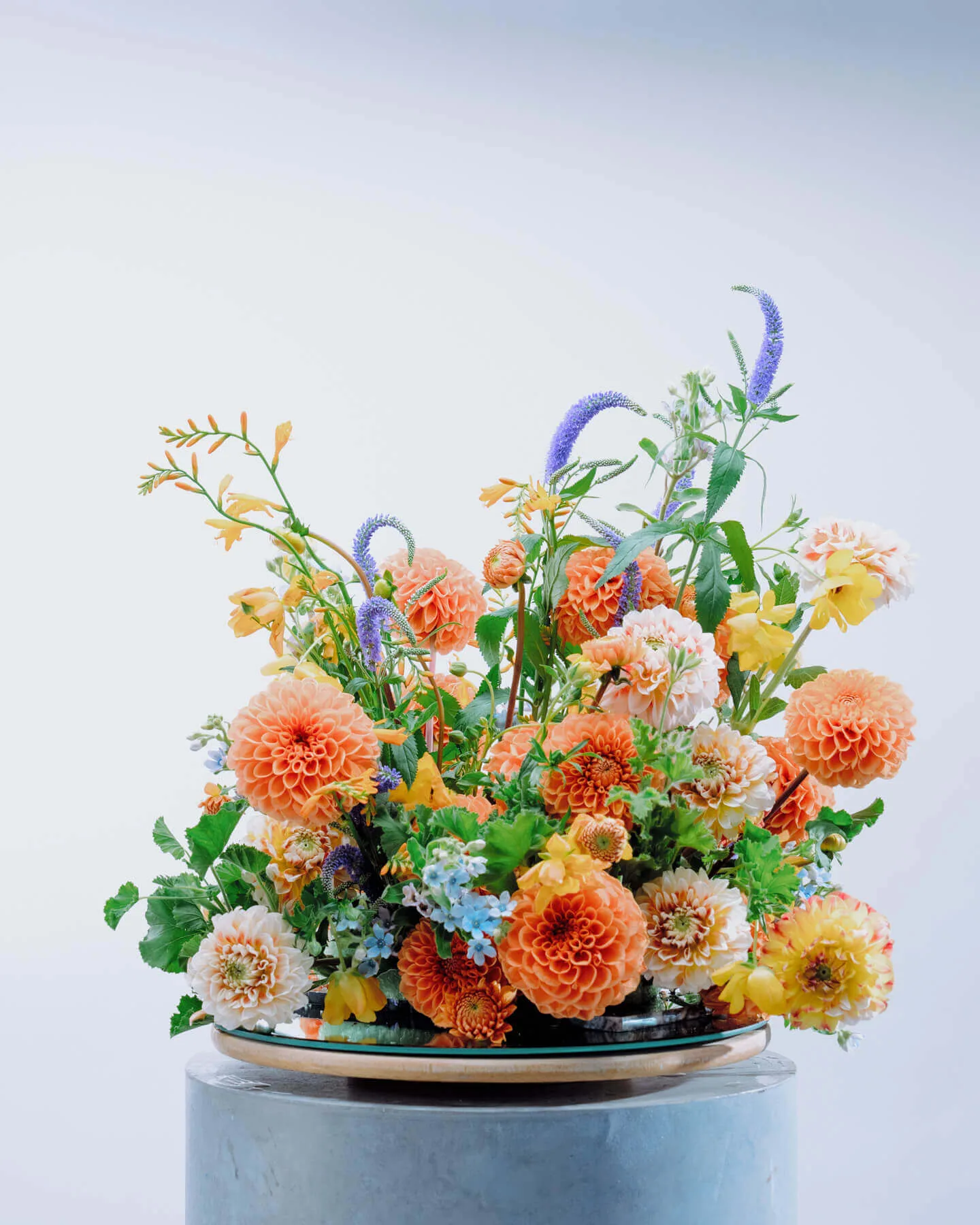 A vibrant floral arrangement on a round grey pedestal with a base of floral foam, featuring orange dahlias, yellow and white flowers, and purple veronica spikes