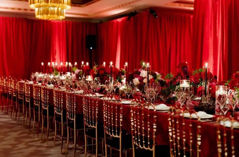 Long table set for an event with deep red flower centrepieces and a background of deep red curtains