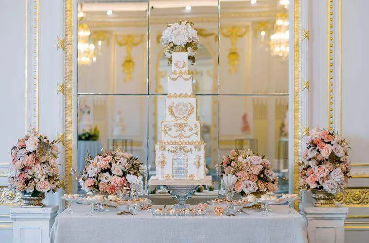A tall wedding cake with floral table displays on either side by blooming haus
