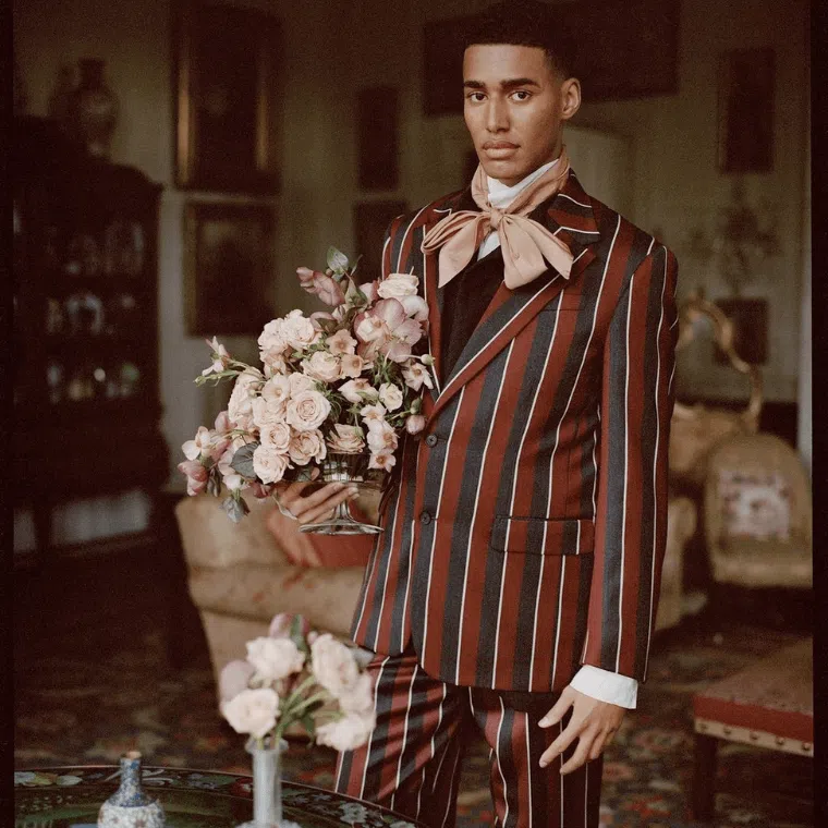 A man dressed in a striped suit holding a pale pink rose bouquet with a backdrop of antique furnishings