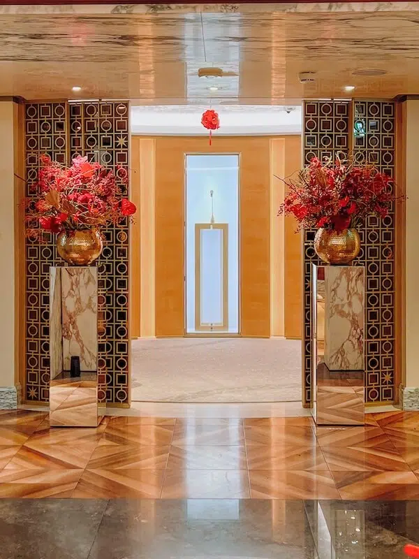 Elegant interior with a decorative entrance featuring two large vases of red flowers, expertly arranged by a renowned London contract florist, on marble pedestals. The doorway is framed with patterned black and gold tiles. A light wood-finished wall and a central display with soft lighting are visible in the background.