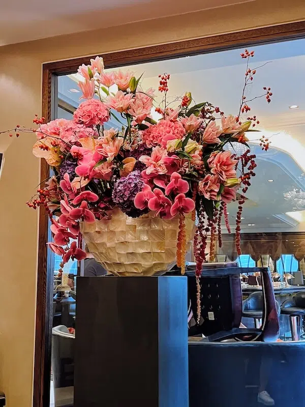 A large, ornate floral arrangement by a London contract florist in a textured white vase sits on a tall black pedestal. Pink lilies, roses, hydrangeas, calla lilies, and cascading red berries are enhanced by soft lighting against the reflective glass backdrop, showcasing its vibrant colors.