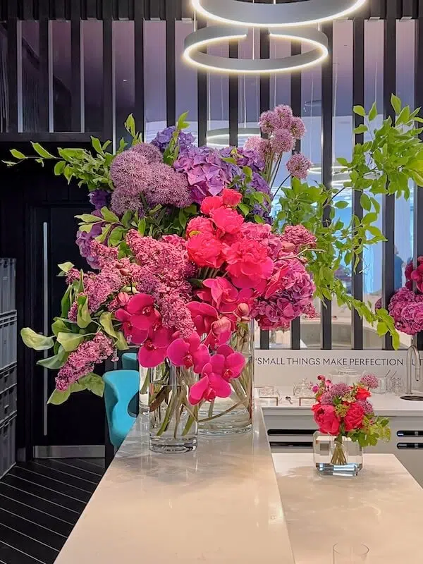 A bouquet of vibrant pink, purple, and green flowers in a clear vase graces a white countertop. Arranged by a London contract florist, the display includes orchids, hydrangeas, and lush greenery. A modern light fixture hangs above against dark vertical panels. A chair is partially visible.