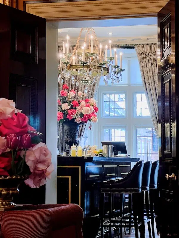 A luxurious room features a bar with high stools, a large ornate chandelier, and a vase of vibrant pink and white flowers arranged by a renowned London contract florist. Tall windows allow natural light to illuminate the space, highlighting the elegant decor. A second bouquet of roses enhances the opulent ambiance.