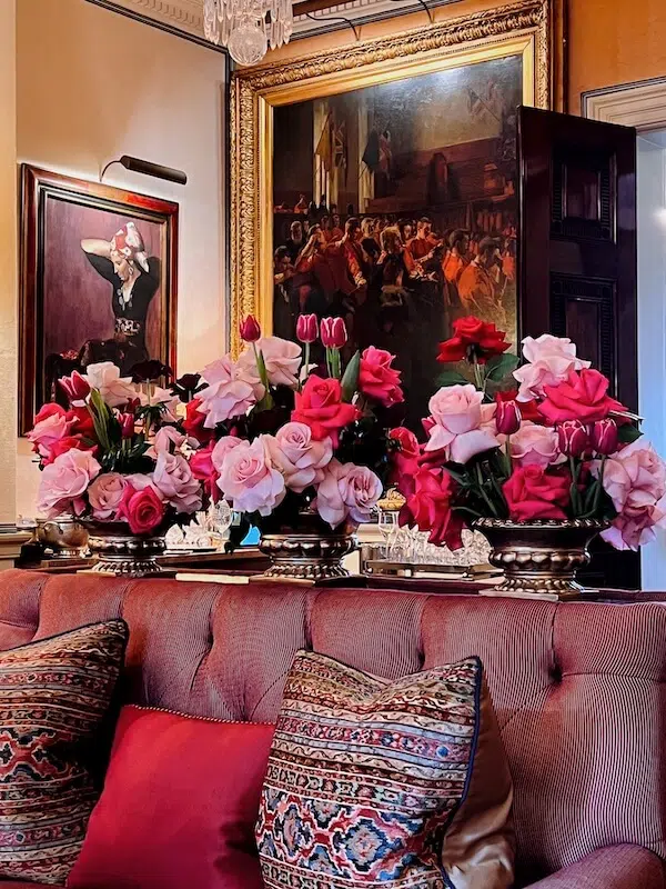 A luxurious room features elegant flower arrangements, courtesy of a renowned London contract florist, with pink and red roses and tulips on a tufted velvet sofa. Decorative pillows with intricate patterns complement the scene, while ornate framed paintings adorn the walls, enhancing the opulent setting.