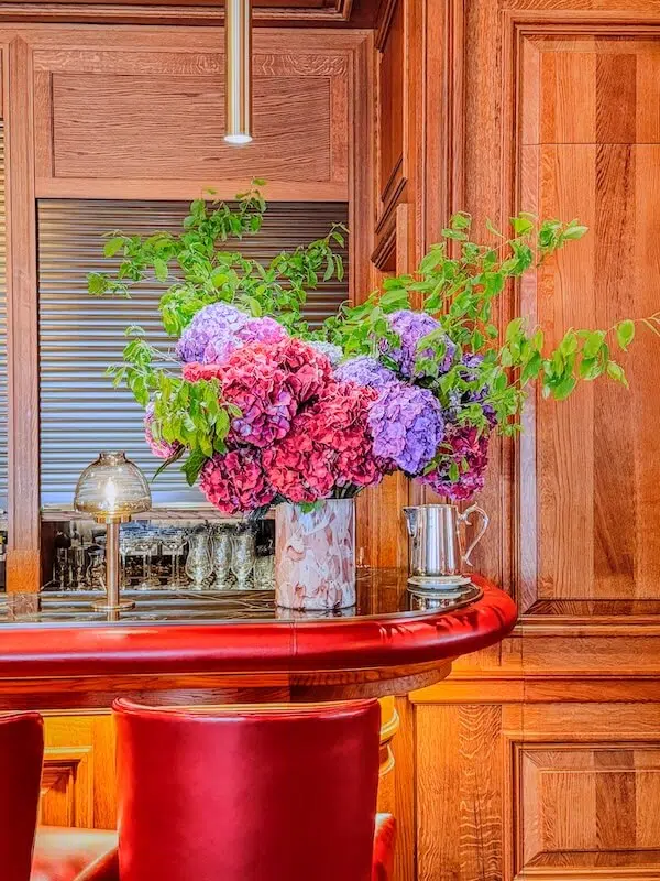 A luxurious bar setting with a red leather counter and chairs, featuring contract flowers: a large bouquet of vibrant purple and pink hydrangeas with green leaves. The wooden walls and a metallic lamp create a warm, elegant ambiance.