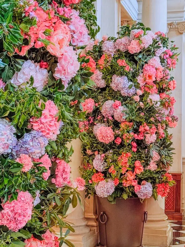 Two large floral arrangements featuring pink and purple hydrangeas and various pink flowers are set against white columns. The lush, cascading blooms showcase the expertise of a London contract florist, creating a vibrant and luxurious indoor garden atmosphere.