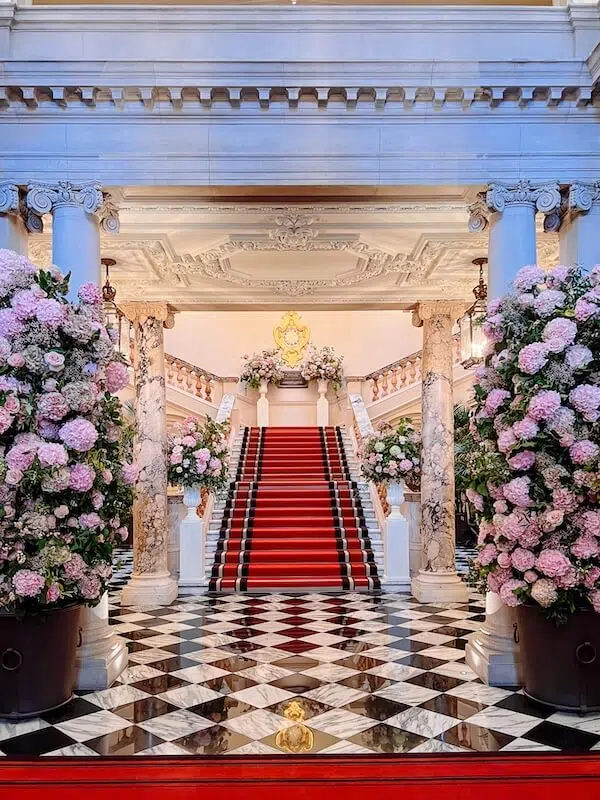 Grand interior with a wide, red-carpeted staircase flanked by ornate white railings. Marble columns and lush arrangements of pink hydrangeas, expertly curated by a renowned London contract florist, adorn both sides. The floor's black-and-white checkered pattern and the ornate ceiling add elegance to the luxurious setting.