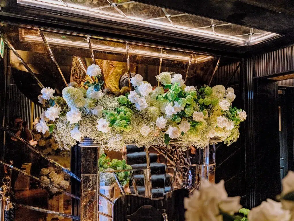 A luxurious floral arrangement by a renowned London contract florist adorns a mirrored mantelpiece, featuring white roses and green hydrangeas mixed with delicate baby's breath. The opulent setting includes reflective surfaces and elegant lighting, enhancing the display's grandeur in a sophisticated room.