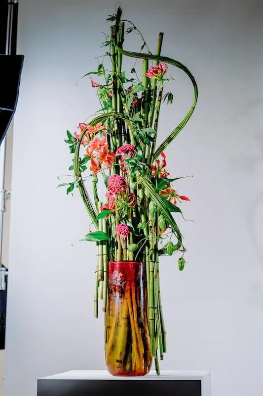 A tall, artistic floral arrangement from a renowned London contract florist is set in a textured, tall amber vase. The arrangement features lush greenery, including vertical bamboo stalks and swirling vines, interspersed with vibrant pink and orange flowers. The plain light gray background highlights the display.