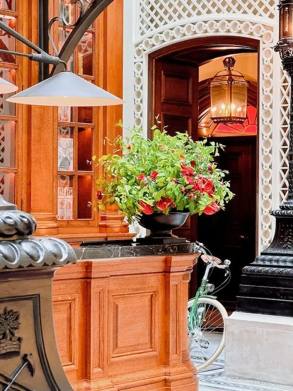 A lavish interior features a wooden desk adorned with contract flowers by a London contract florist, showcasing a vibrant bouquet of red and green blooms in a black vase. Behind, an intricate lattice archway and wooden door add charm, while elegant lighting casts a warm glow on the mint-green bicycle frame.