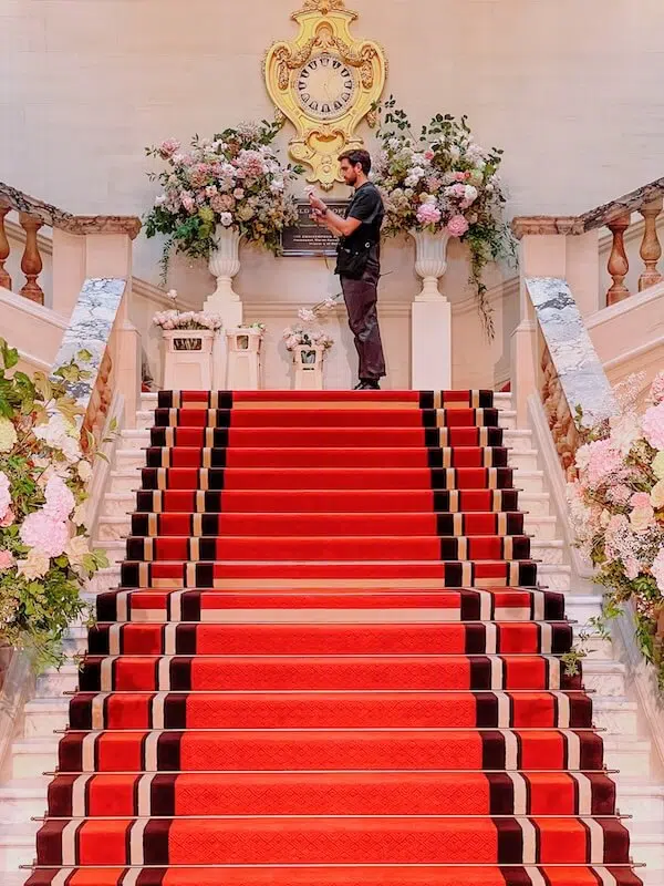 A man stands on a grand red-carpeted staircase, photographing with a camera. The elegant staircase, courtesy of a top London contract florist, is lined with marble banisters and adorned with lush floral arrangements. A large ornate clock is mounted on the wall above, surrounded by decorative wall sconces and plaques.