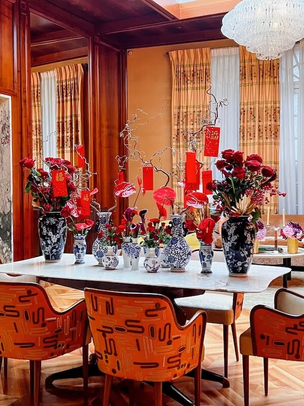 A dining room in London showcases a white marble table adorned by exquisite arrangements from a contract florist, with blue and white vases filled with red flowers and decorative branches. Red lanterns hang amid patterned orange chairs, wooden walls, curtains, and a grand chandelier.