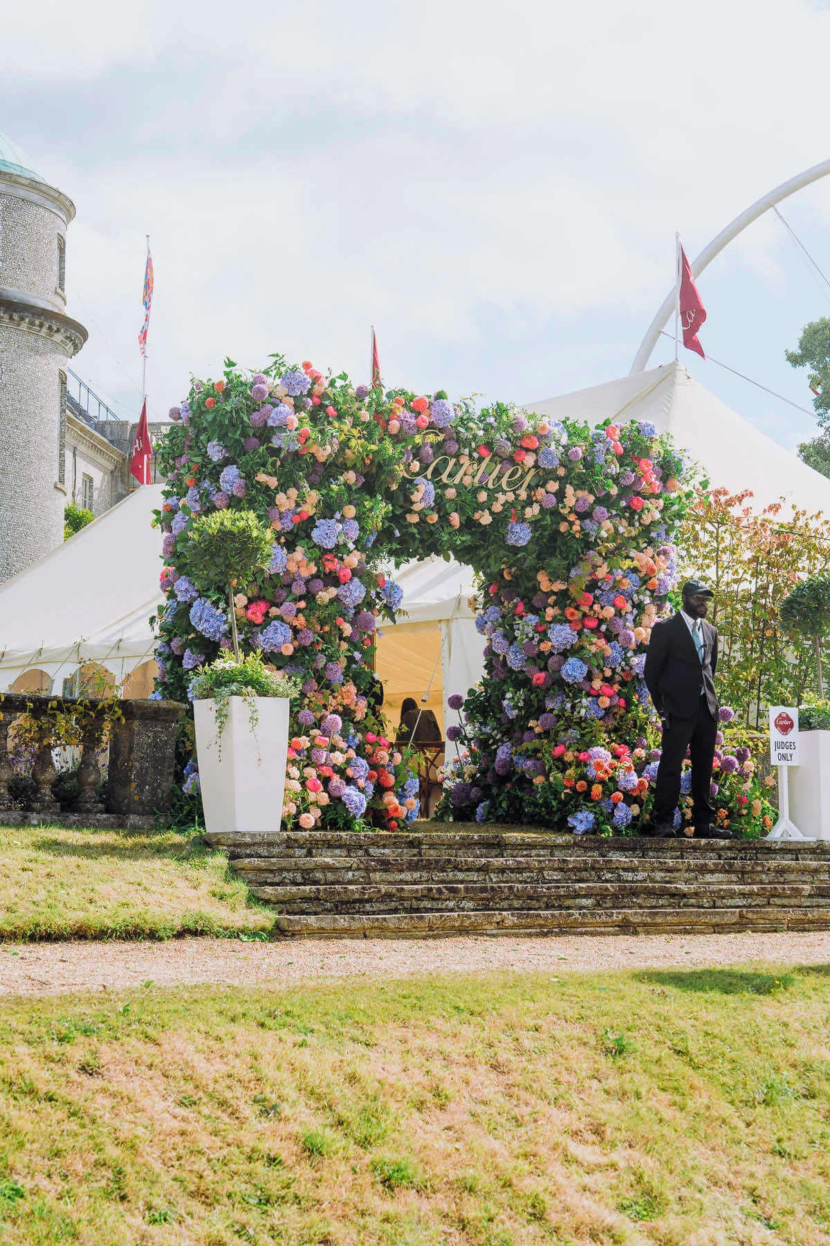 A large, flower-adorned archway made by Blooming Haus at the entrance of a white event tent. The arch is covered in colorful hydrangeas and roses. A suited man stands near a sign reading "Cartier." The tent is set up in front of a historic building with multiple red flags flying in the background.