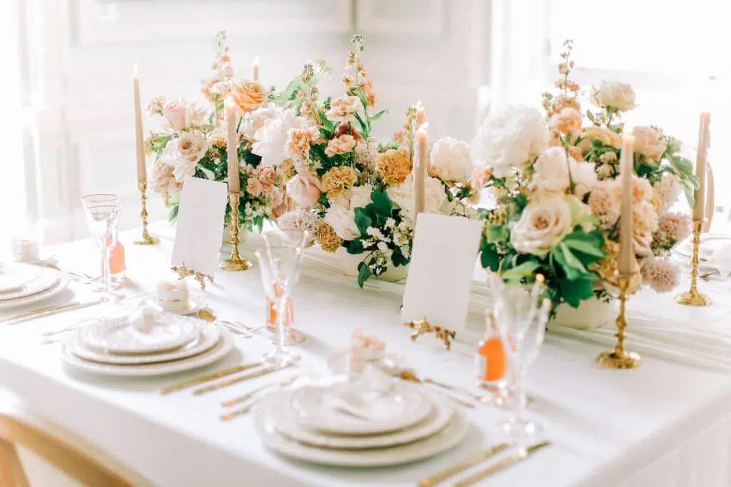 A beautifully set dining table with a white tablecloth, adorned with elegant floral arrangements by a luxury florist featuring pastel roses and greenery. Gold candlesticks with pale pink candles are interspersed among the flowers. White plates, gold utensils, and crystal glasses are neatly arranged, with name cards at each setting.