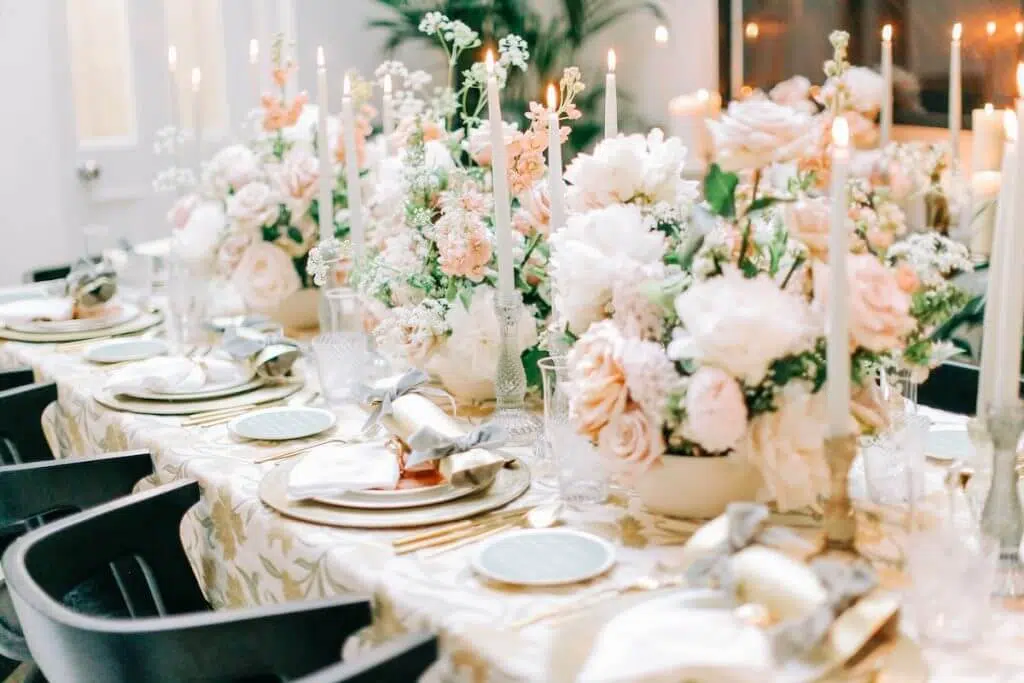 An elegantly set dining table adorned with a white and gold patterned tablecloth, featuring lush bouquets of white and blush pink flowers from a luxury London florist. Tall, slender white candles in gold holders are interspersed among the floral arrangements. Each place setting includes fine china, crystal glassware, and neatly folded napkins.
