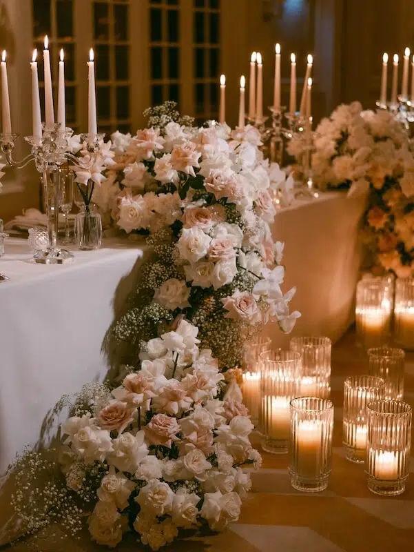 A lavish wedding reception table adorned with white and blush pink roses, flanked by tall, lit candles in glass holders. The table is draped in a white cloth, with cascading floral arrangements along the edge from the finest London florist, creating a romantic and elegant ambiance.
