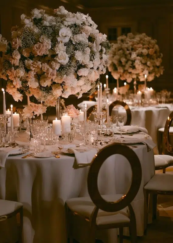 A sophisticated wedding reception setup designed by a luxury London florist features round tables adorned with lush, tall floral centerpieces of white and blush roses mixed with greenery. Each table is elegantly set with white linens, fine glassware, and lit candles. Stylish gold chairs with circular backs surround the tables.