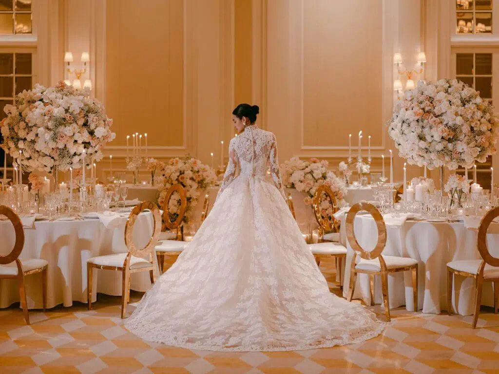 A bride in an elegant, long-sleeved lace wedding gown stands with her back to the camera in a lavishly decorated reception hall. The room, styled by a top London florist, features round tables adorned with tall floral centerpieces and lit candles, along with gold-accented chairs, creating a romantic and sophisticated ambiance.