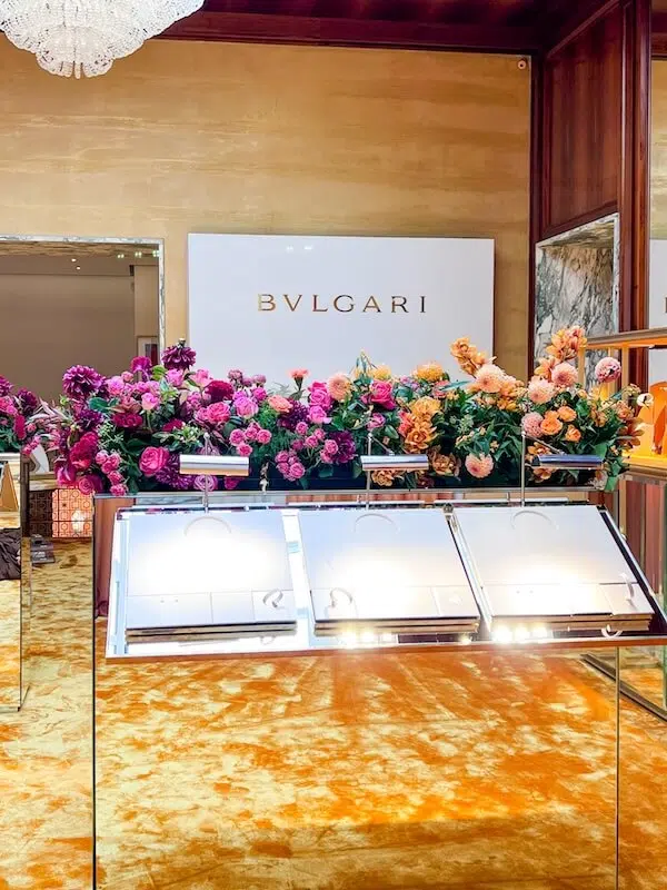 A luxurious Bvlgari store interior features a gold carpet, mirrored display cases, and vibrant contract flowers arranged with pink, purple, and orange blooms. The Bvlgari logo graces a white wall, while a chandelier above elevates the elegance—a testament to London's renowned contract florist.