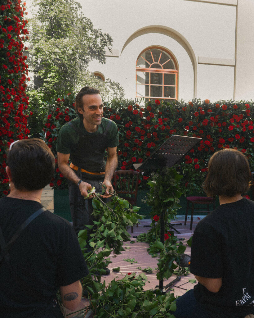 Michael, Blooming Haus's Operations Director, in a green shirt and pants stands on a small outdoor stage decorated with foliage, leaning forward and smiling. He holds a leafy branch in his hands. Behind him is a tall red rose wall from the event florist, and a building with arched windows. Two people with short hair sit facing him.