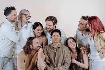 A group of nine Blooming Haus Team members stand closely together, smiling and interacting warmly. Casually dressed in neutral tones, they create a harmonious look. The plain off-white background keeps the focus on their happy expressions and relaxed poses.
