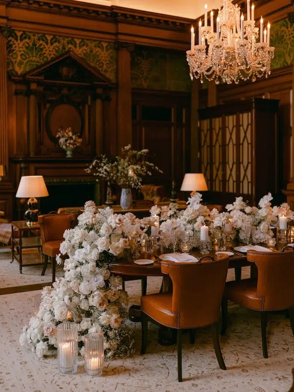 An opulent dining room features a long wooden table adorned with an abundance of event flowers cascading over the edge. Ornate chairs surround the table, and crystal chandeliers hang from a high ceiling. Elegant lamps and a detailed fireplace enhance the luxurious atmosphere curated by a luxury London florist.