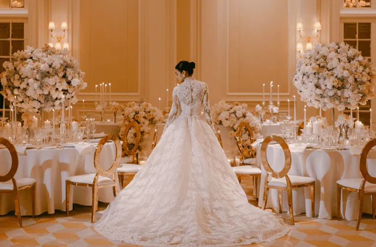 A bride in an elegant, long-sleeved lace wedding gown stands in the center of an opulently decorated reception hall reminiscent of a bridgerton ball. Round tables with white tablecloths are adorned with large, lush floral centerpieces and surrounded by gold chairs. Candles and soft lighting create a romantic atmosphere.