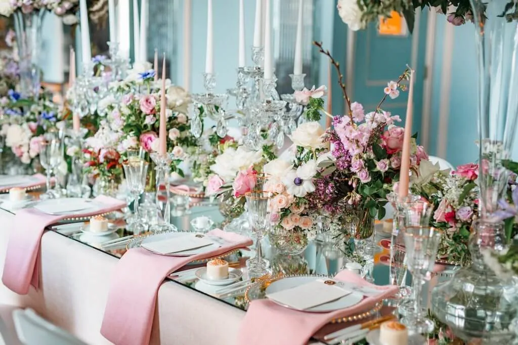 A lavishly set dining table decorated with exquisite wedding flowers in shades of pink, white, and purple. Elegant glassware, candles, and gold-rimmed plates are arranged on the mirrored surface. Pink napkins are neatly placed on the plates, creating a sophisticated and romantic ambiance.
