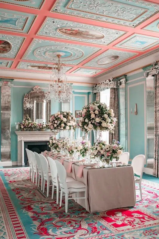 An elegantly decorated dining room features a long table set for a formal meal. Adorned with wedding flowers by a luxury London florist, the table boasts a beige tablecloth, tall flower arrangements, and white chairs. The room has ornate pastel-colored ceiling details, a chandelier, large mirror above the fireplace, and floral-patterned carpet.