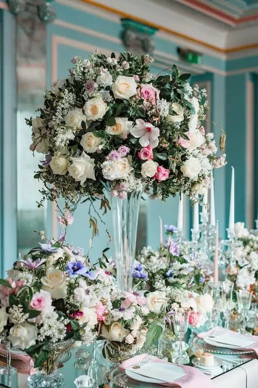 A lavish floral arrangement adorns an elegantly set dining table, featuring a tall, clear vase brimming with wedding flowers in white, pink, and purple roses, and assorted greenery. Additional smaller bouquets from a London florist, candles, and glassware complement the pastel-themed decor in an opulent room with blue and white walls.