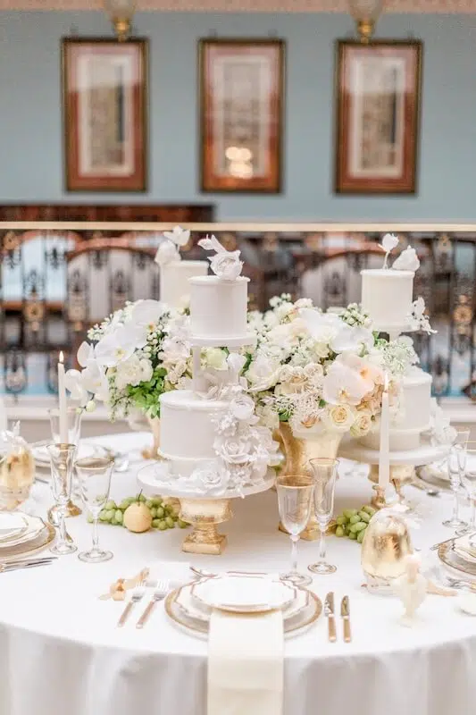An elegantly set table features a white tablecloth, gold-rimmed plates, silverware, and tall candles. Centerpieces include white floral arrangements from a luxury London florist with roses and hydrangeas. Multiple tiered cakes adorned with white flowers, grapes, and gold-colored details complete the decor.