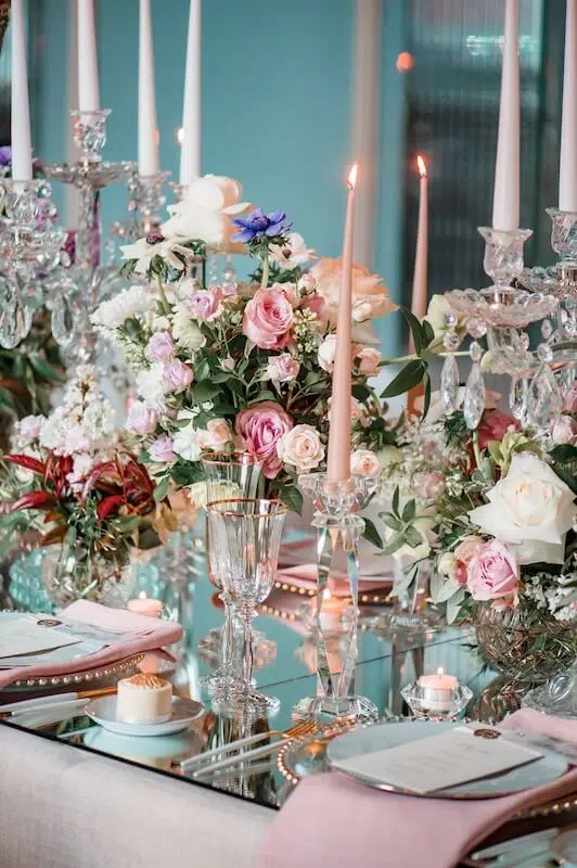 A lavishly set table features elegant wedding flowers in pink, white, and purple arrangements, crystal candle holders with lit tapered candles, and delicate porcelain plates with pink napkins. Glasses and silverware are neatly arranged on a reflective, mirrored surface.