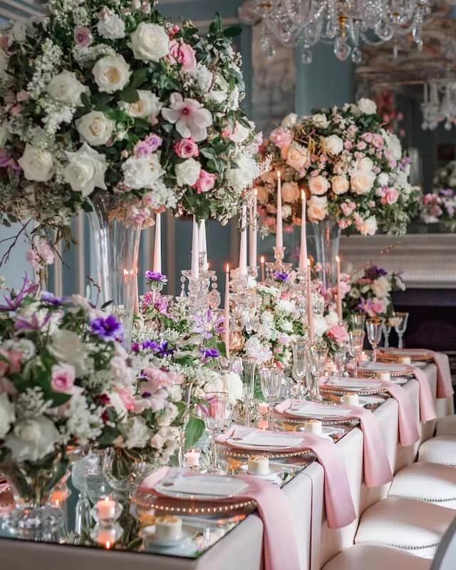 A lavishly decorated dining table is set for a formal event. It is adorned with tall floral centerpieces of wedding flowers in white, pink, and purple. Long, white candles in crystal holders are lit, adding a warm glow. Pink table runners, elegant plates, and glassware complete the sophisticated look from a luxury florist.