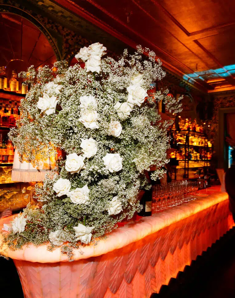 A lavish bar counter adorned with a flowing arrangement of wedding flowers, including white roses and baby's breath. Behind the bar, shelves stocked with bottles of liquor are illuminated warmly. The ceiling and walls feature ornate, intricate designs adding to the opulent atmosphere. Glassware is neatly arranged on the counter.