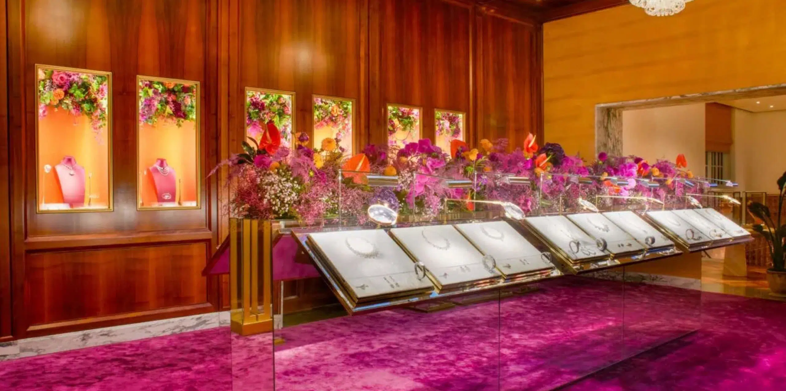 The image shows an ornate jewelry display in a luxurious setting. The jewelry pieces are showcased in glass cases, surrounded by vibrant floral arrangements from a luxury London florist, featuring purple, pink, and red flowers. The wooden walls and plush purple carpet enhance the opulent atmosphere.
