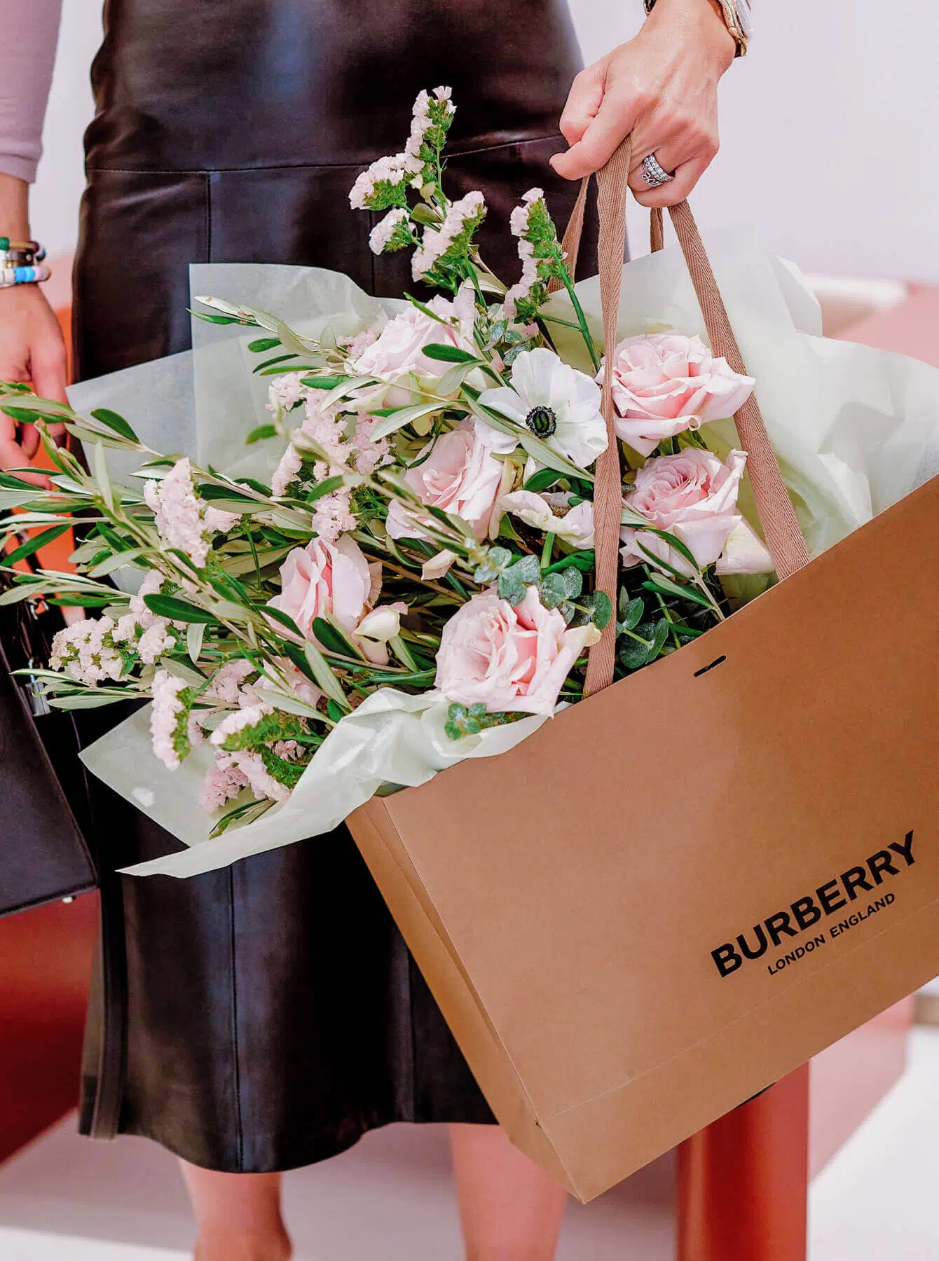 A person in a dark leather skirt holds a brown Burberry shopping bag filled with pink and white flowers and greenery from a luxury florist. Another bag is partially visible in the background, suggesting a chic, fashionable setting with an emphasis on style and elegance, reminiscent of London flowers.