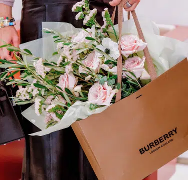 A person holds a Burberry paper shopping bag filled with a bouquet of pink roses and white flowers from a luxury London florist wrapped in white paper. The person is also carrying a black handbag and wearing several colorful bracelets. The background is blurred, focusing on the London flowers and the bag.