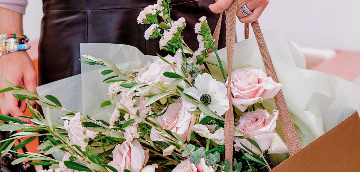 A person in a dark skirt holds a paper bag containing a bouquet of pale pink roses, white wedding flowers, and green foliage. Their hand, adorned with a ring and colorful bracelets, grasps the bag's handles. The bouquet, arranged by a luxury London florist, is wrapped in soft green tissue paper.