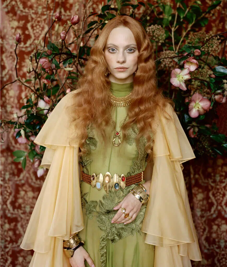 A person with long red hair stands against a backdrop of editorial flowers, exuding an aura of elegance. They are wearing a flowing yellow cape over a green dress adorned with embroidery. The detailed belt, embellished with stones and ornate jewelry, perfectly complements the lush floral styling surrounding them.