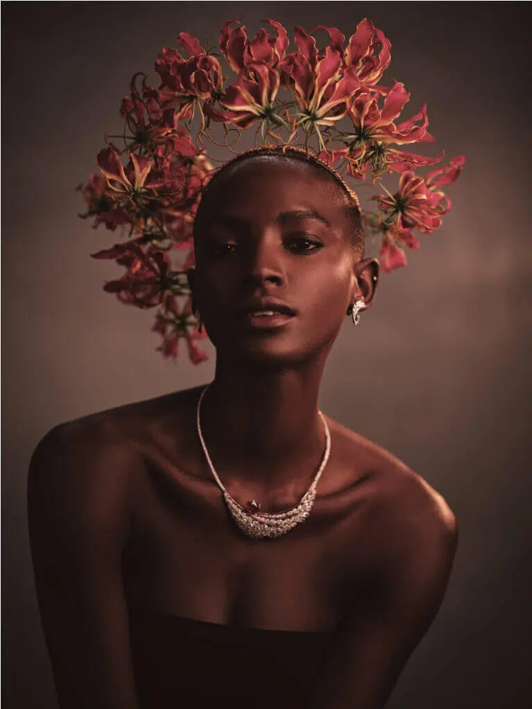 A person poses against a dark background, wearing a crown crafted with editorial flowers. They have short hair and are adorned with sparkling earrings and a necklace. Their strapless clothing emphasizes their shoulders and collarbone, while the lighting casts soft shadows, adding drama to this floral styling scene.
