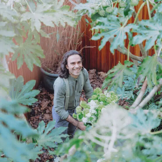 A person with long hair, wearing a green shirt and suspenders, is standing in a lush garden surrounded by large green leaves. They are smiling and holding a bouquet of green and white flowers. The background features a wooden fence and potted plants, highlighting the serene beauty of sustainable living.