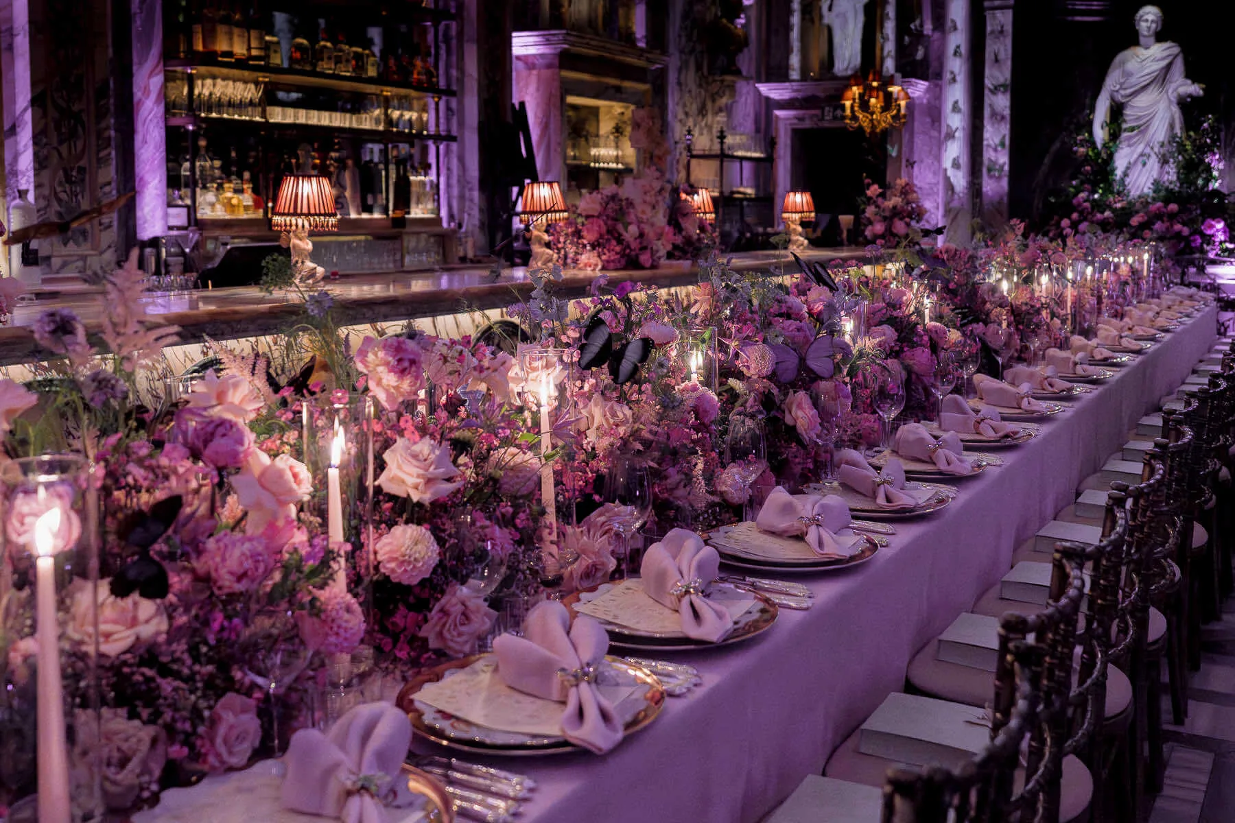 An elegantly set long dining table with intricate floral arrangements in shades of purple and pink, masterfully arranged by a corporate florist. The table features white napkins, gold-rimmed plates, and lit candles. Behind, there are shelves lined with bottles, a statue, and purple ambient lighting, creating a luxurious atmosphere. Photo taken in Bacchanalia, London.
