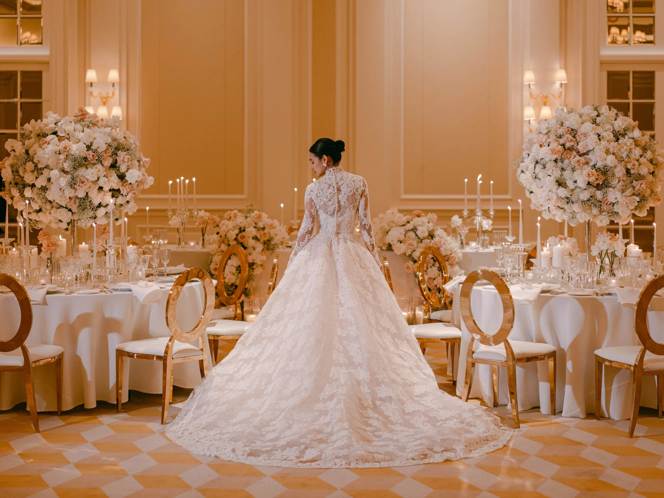 A bride in a lace wedding gown stands with her back to the camera in an elegant, softly lit ballroom. The room, styled by a luxury florist, features round tables with tall floral centerpieces, candles, and gold-accented chairs. The floor showcases a geometric pattern in cream and white tones.