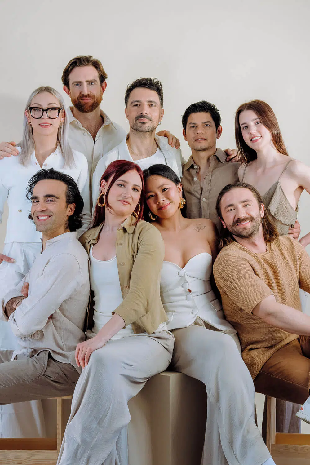 A group of nine people is posing together in front of a beige background, dressed in neutral tones of white, beige, and brown. With warm expressions and casual poses, they resemble a luxury florist team from London, capturing both elegance and closeness.