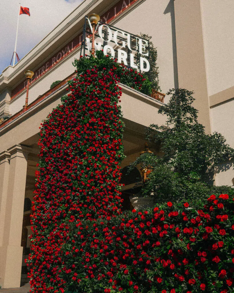 Theatre entrance adorned with vibrant red roses, courtesy of a luxury florist, climbing up archways and walls. A sign with the words "Vogue World" is perched atop the building. Flanked by large columns in classic architectural style, a flag waves at the top against a partly cloudy sky.