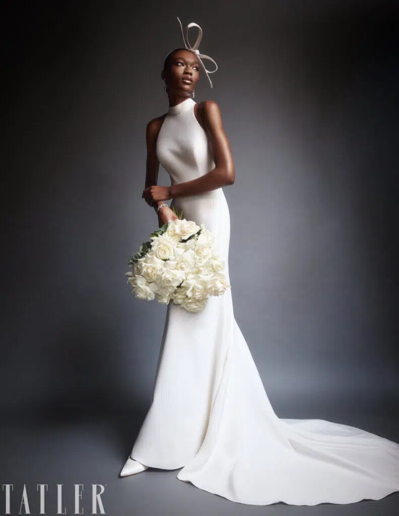 A woman in a sleek, form-fitting white gown poses gracefully against a dark background, showcasing exquisite editorial flowers. She holds a large bouquet of white roses. Her ensemble includes a unique headpiece with a looped design. The dress has a long train, and she wears pointed white shoes. "TATLER" is visible.