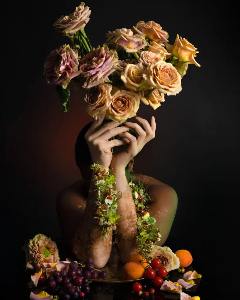 A person obscured by a bouquet of cream and pink roses held over their face, showcasing exquisite floral styling. Their arms are adorned with vines and leaves. Grapes, apples, and other fruits gather in the foreground, while the dark backdrop highlights the vibrant editorial flowers and fruits.