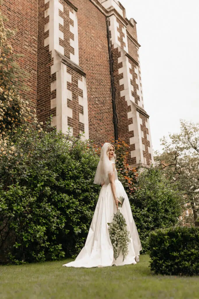 Bride in a white gown and veil holds a bouquet of greenery with exquisite floral styling, standing on grass in front of a tall brick building with geometric patterns. She is gazing upwards, surrounded by lush bushes and trees, beneath a cloudy sky.