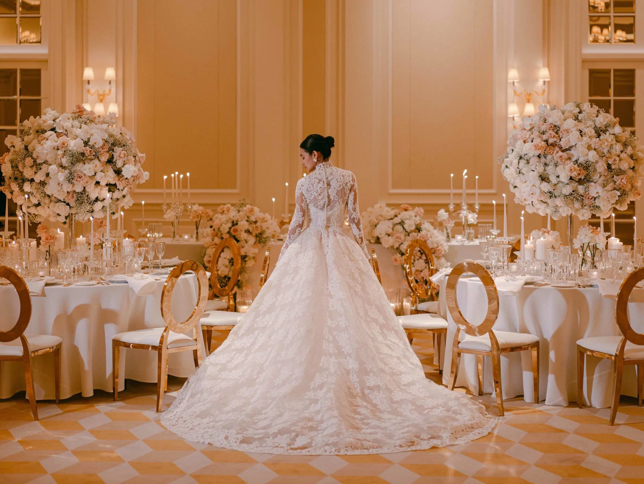 A bride in an elegant, long-sleeved lace gown stands with her back to the camera in a lavishly decorated ballroom, where a skilled London wedding florist has arranged white and blush floral centerpieces for a romantic, candlelit ambiance. Best wedding flowers in London.